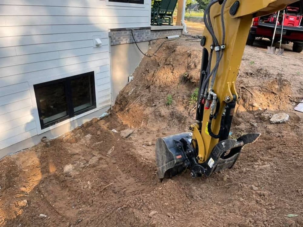 Excavator digging near house foundation for construction or landscaping project.