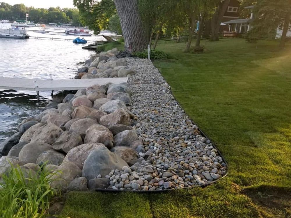 Rocky shoreline with pebbles, grass, and wooden dock by a serene lake setting.